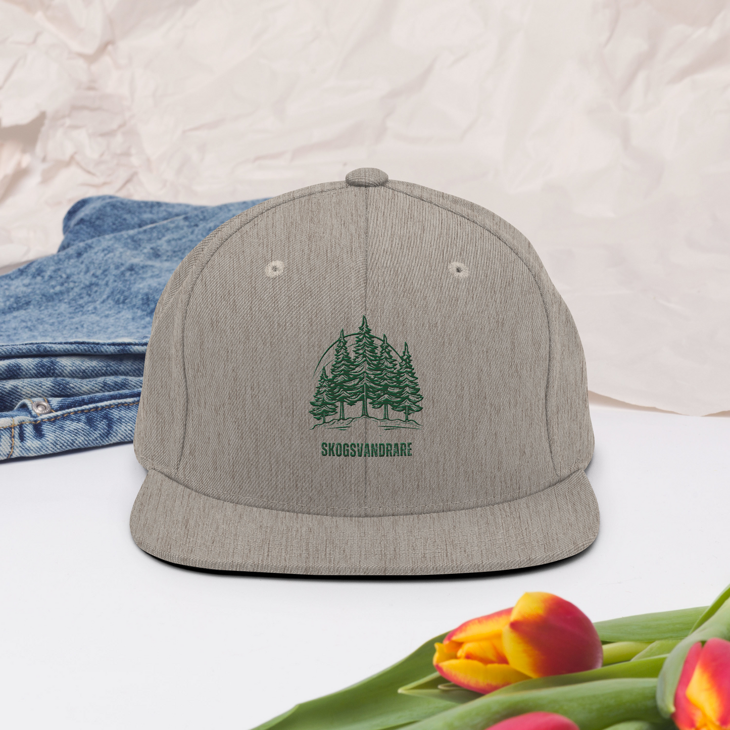Melerad grå snapback keps, broderade granar och texten ”Skogsvandrare” på framsidan.
Heather grey snapback cap, featuring embroidered pine trees and the word “Skogsvandrare” on the front.