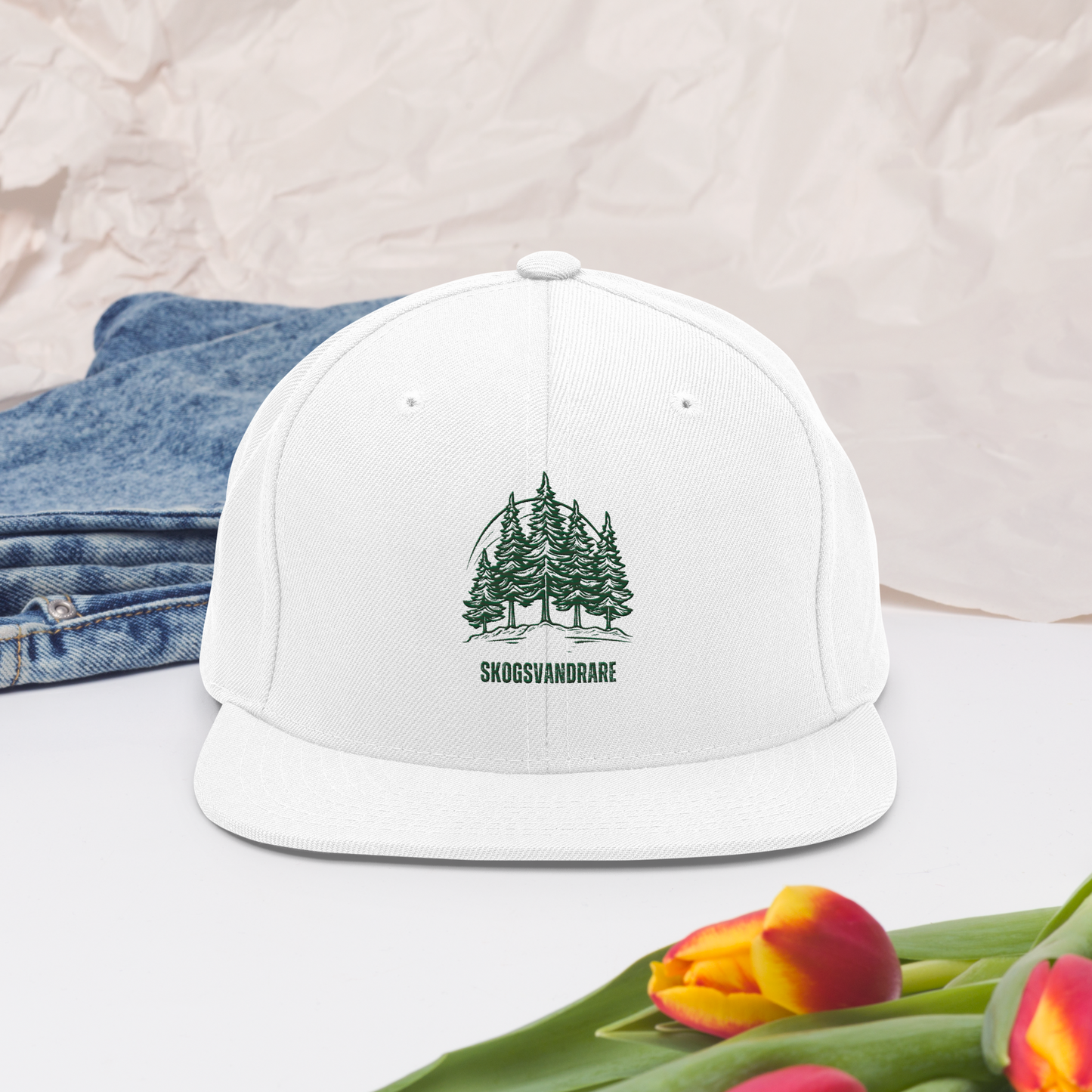 Vit snapback keps, broderade granar och texten ”Skogsvandrare” på framsidan.
White snapback cap, featuring embroidered pine trees and the word “Skogsvandrare” on the front.