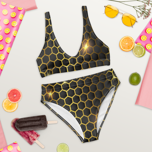 Högmidjad bikini i återvunnet material med glänsande bikakemönster i guld på mörk bakgrund.
High-waisted bikini made from recycled material with a shiny golden honeycomb pattern on a dark background.