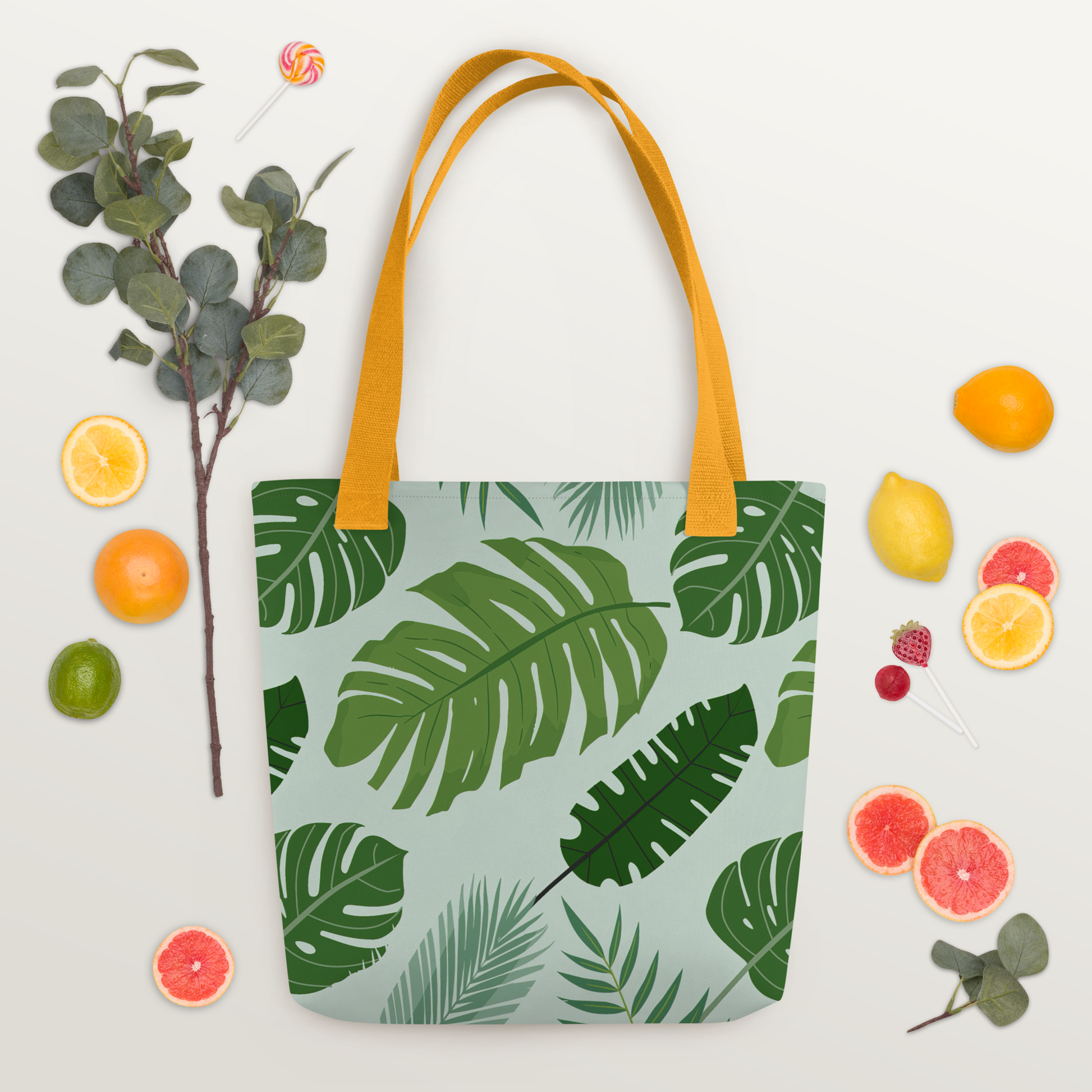 Polyester-tygväska med gult handtag och tropiskt bladmotiv i gröna nyanser på ljus bakgrund.
Polyester tote bag with yellow handle and tropical leaf pattern in green tones on a light background.
