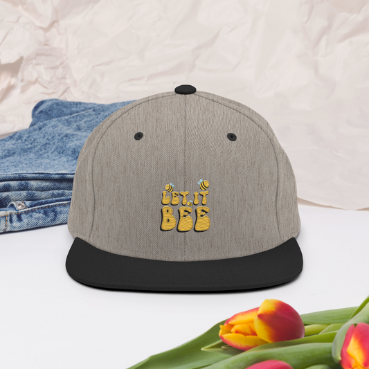 Melerad grå snapback-keps med svart skärm och broderad gul text ”Let it Bee” samt små bin på framsidan.
Heather gray snapback cap with a black brim, featuring embroidered yellow text “Let it Bee” and small bees on the front.