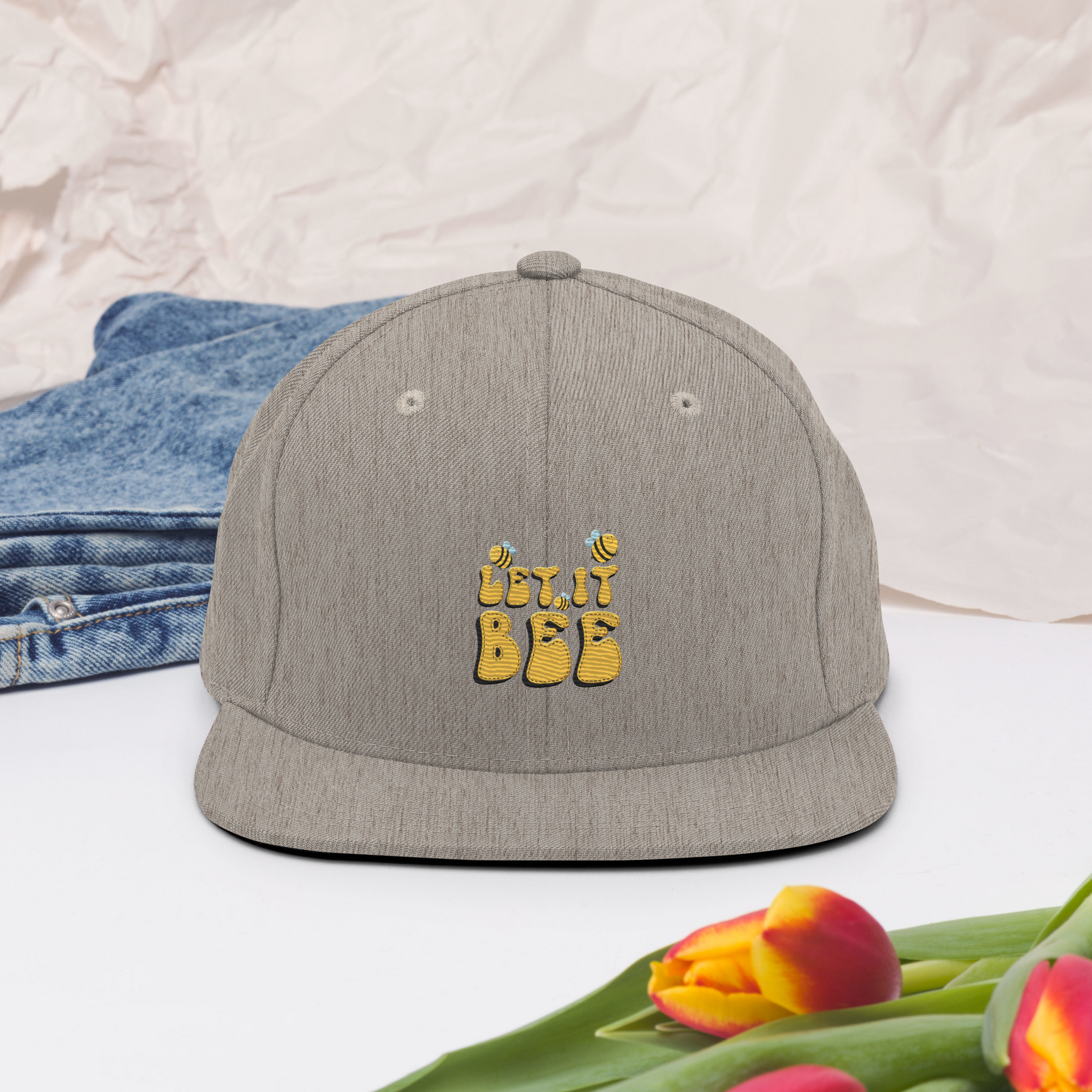 Melerad grå snapback-keps med broderad gul text ”Let it Bee” samt små bin på framsidan.
Heather gray snapback cap, featuring embroidered yellow text “Let it Bee” and small bees on the front.