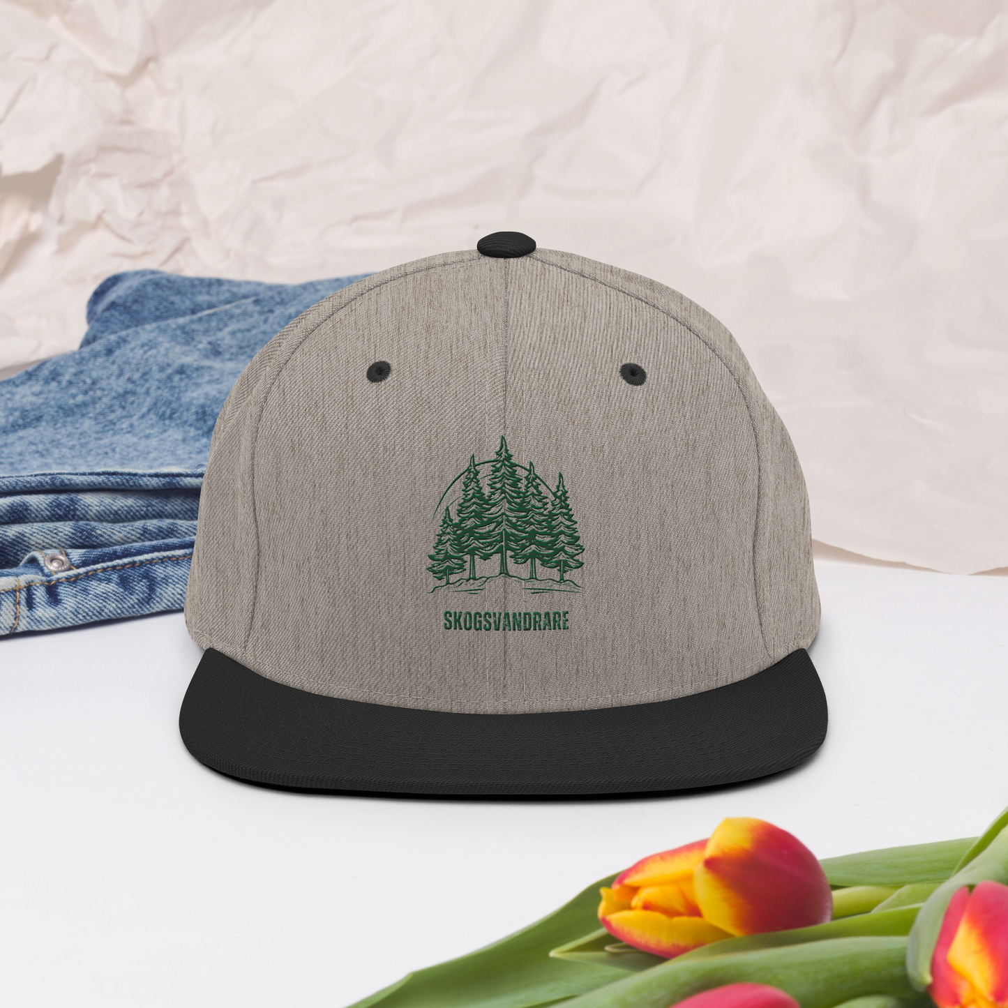 Melerad grå snapback keps med svart skärm, broderade granar och texten ”Skogsvandrare” på framsidan.
Heather grey snapback cap with a black brim, featuring embroidered pine trees and the word “Skogsvandrare” on the front.