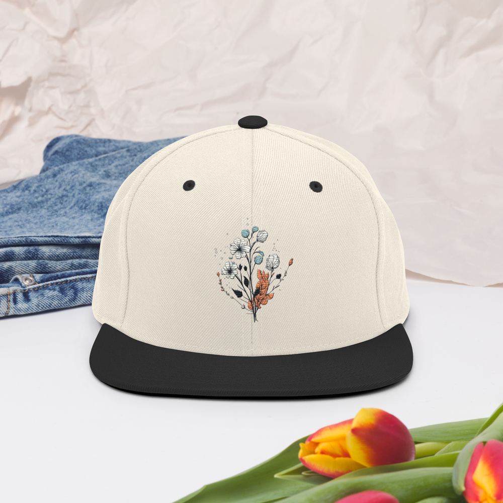Natur snapback keps med svart skärm och broderad blomdesign framtill.
Natural snapback cap with black brim and embroidered floral design on the front.