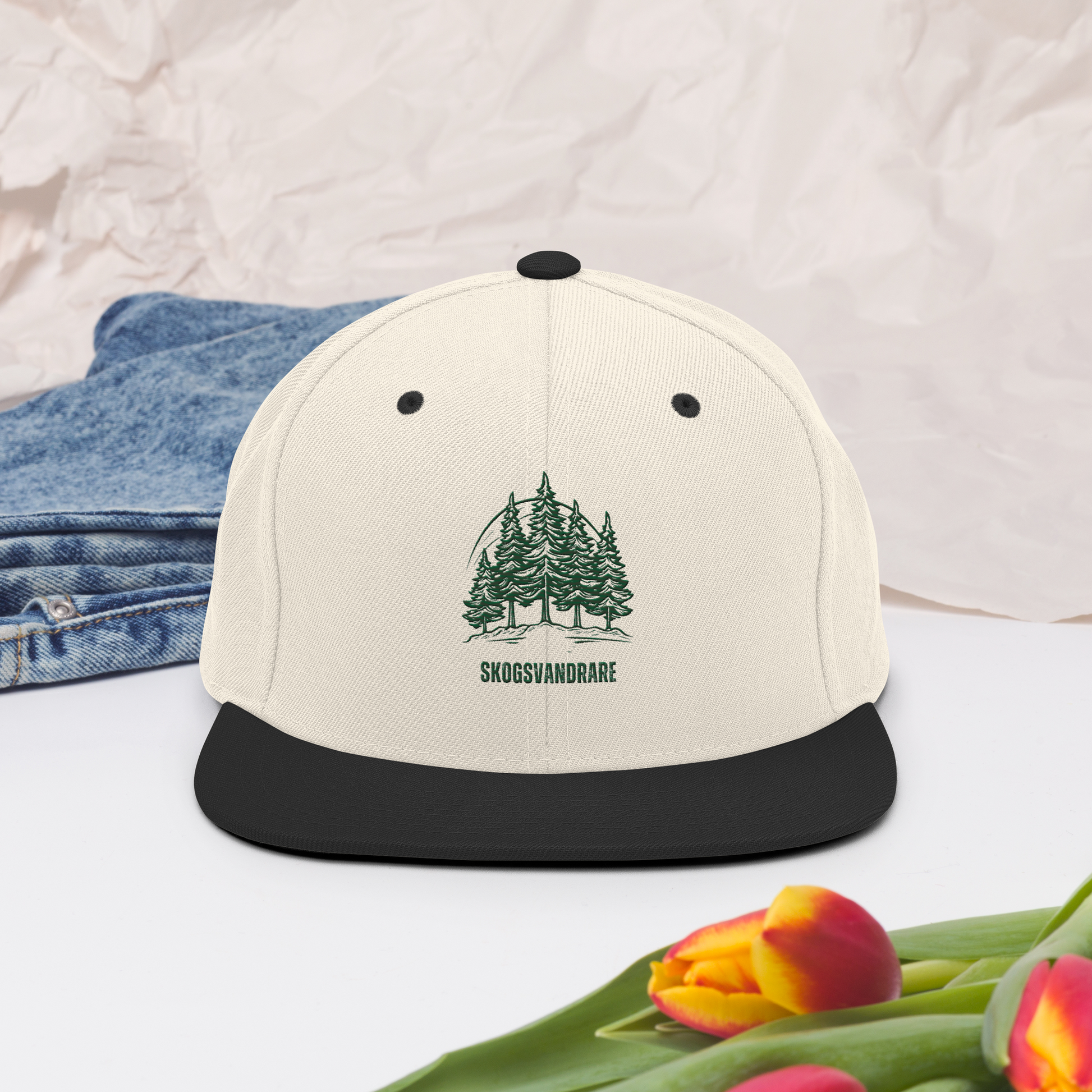 Natur snapback keps med svart skärm, broderade granar och texten ”Skogsvandrare” på framsidan.
Natural snapback cap with a black brim, featuring embroidered pine trees and the word “Skogsvandrare” on the front.