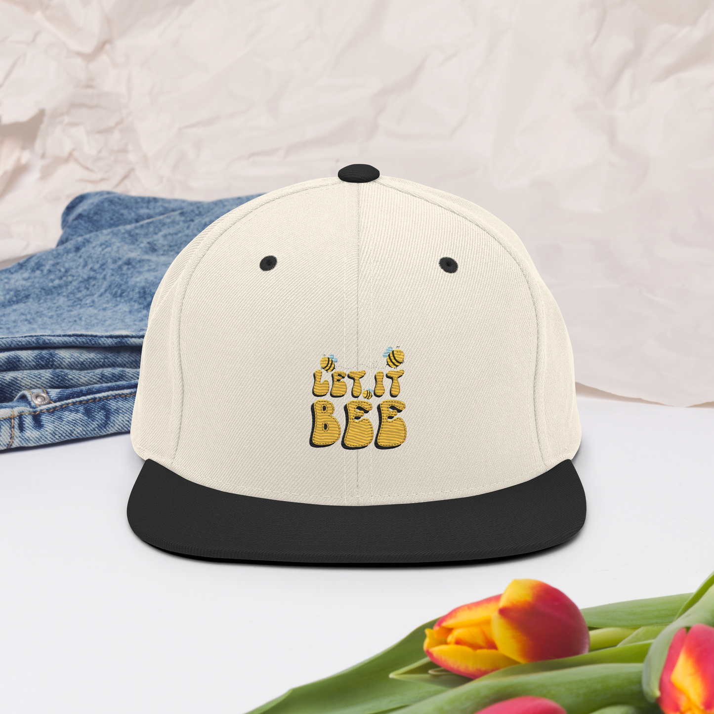Natur snapback-keps med svart skärm och broderad gul text ”Let it Bee” samt små bin på framsidan.
Natural snapback cap with a black brim, featuring embroidered yellow text “Let it Bee” and small bees on the front.