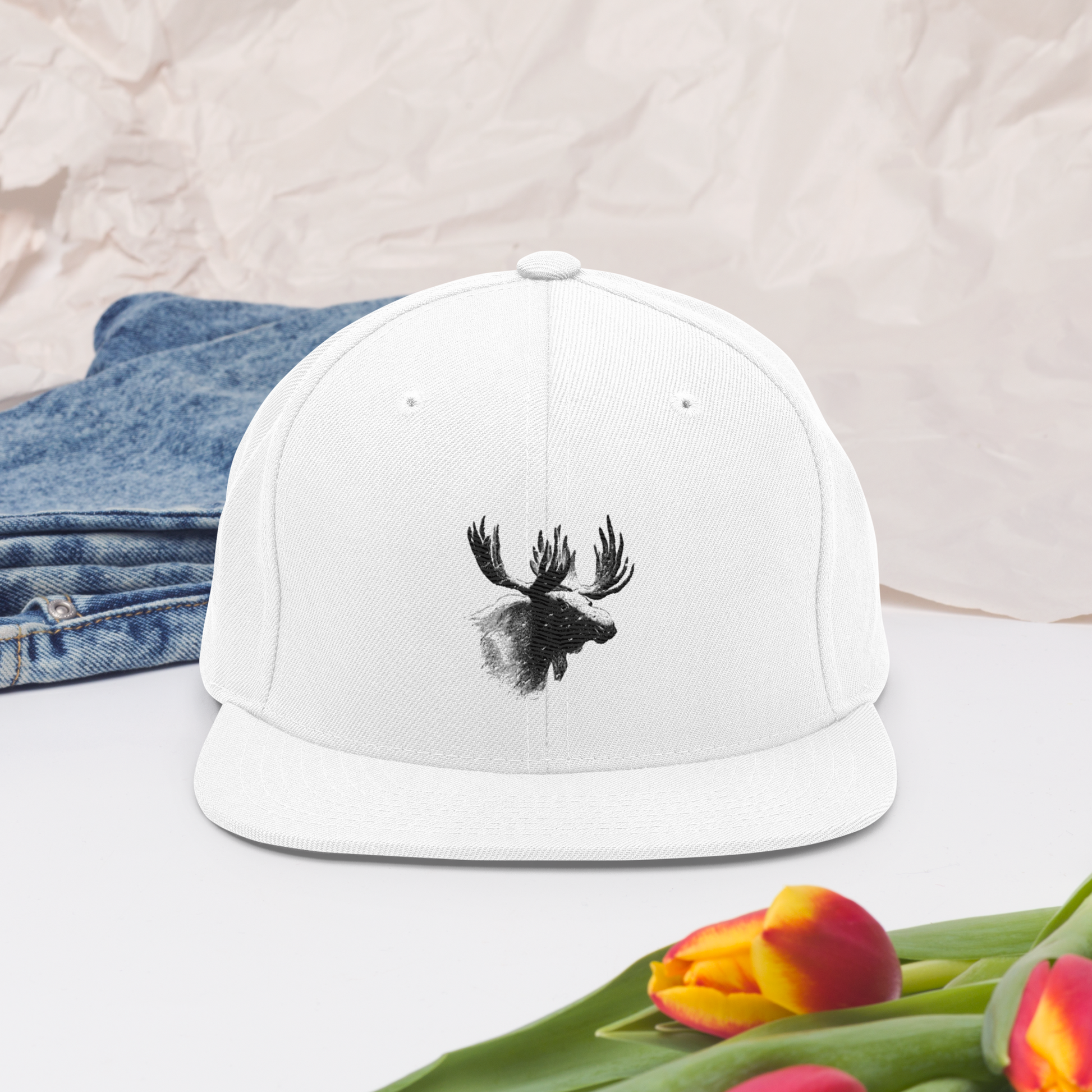Vit snapback-keps med broderat älghuvud i svart på framsidan.
White snapback cap, featuring an embroidered black moose head on the front.