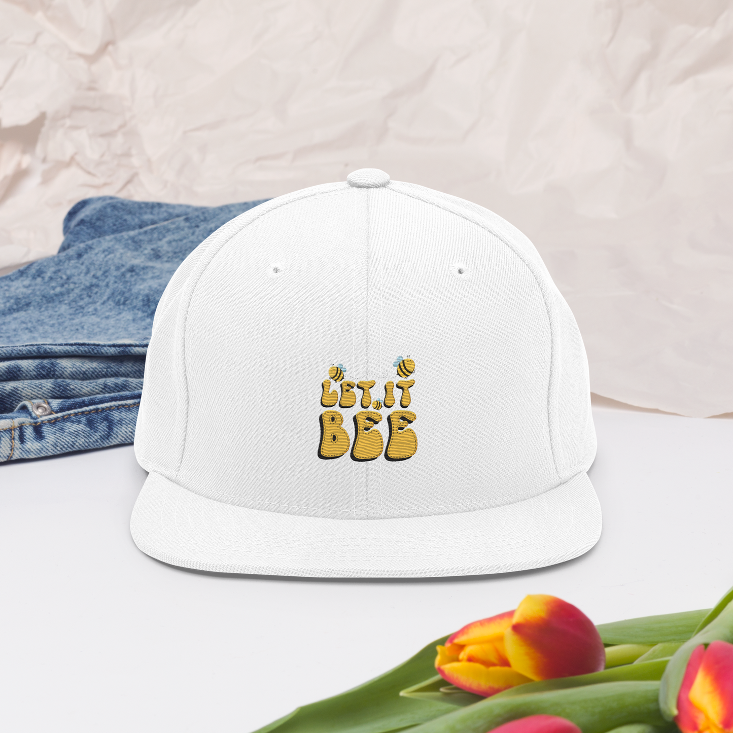 Vit snapback-keps med broderad gul text ”Let it Bee” samt små bin på framsidan.
White snapback cap, featuring embroidered yellow text “Let it Bee” and small bees on the front.