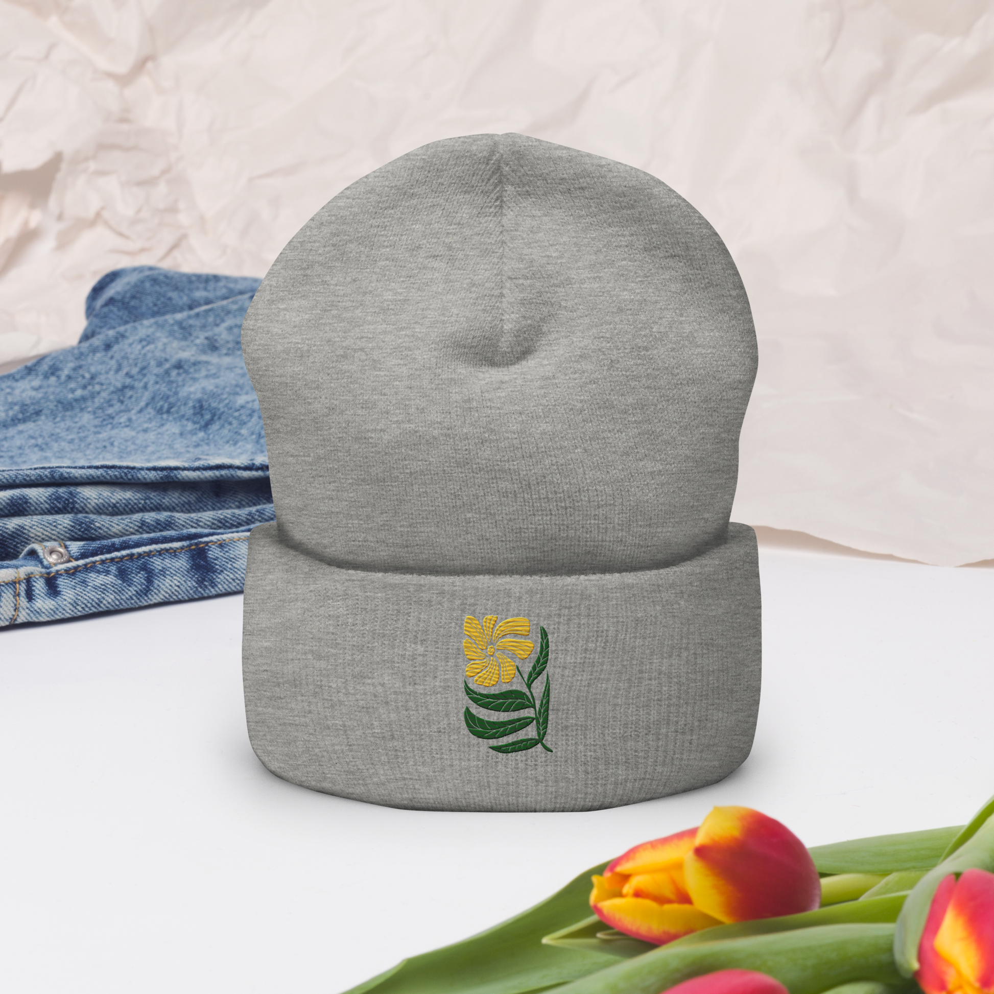 Grå uppvikt mössa med broderad gul blomma och gröna blad framtill.
Grey cuffed beanie with embroidered yellow flower and green leaves on the front.