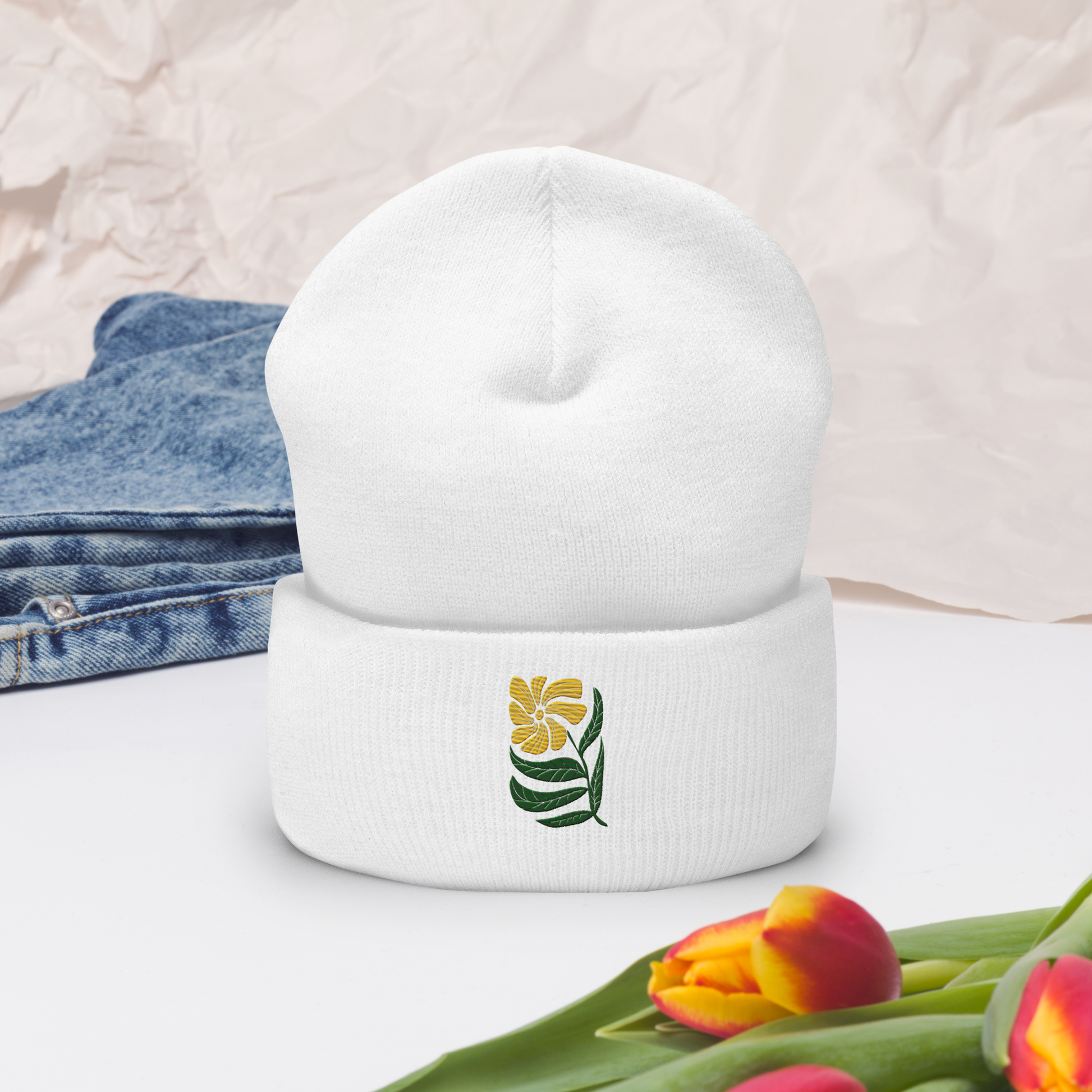 Vit uppvikt mössa med broderad gul blomma och gröna blad framtill.
White cuffed beanie with embroidered yellow flower and green leaves on the front.