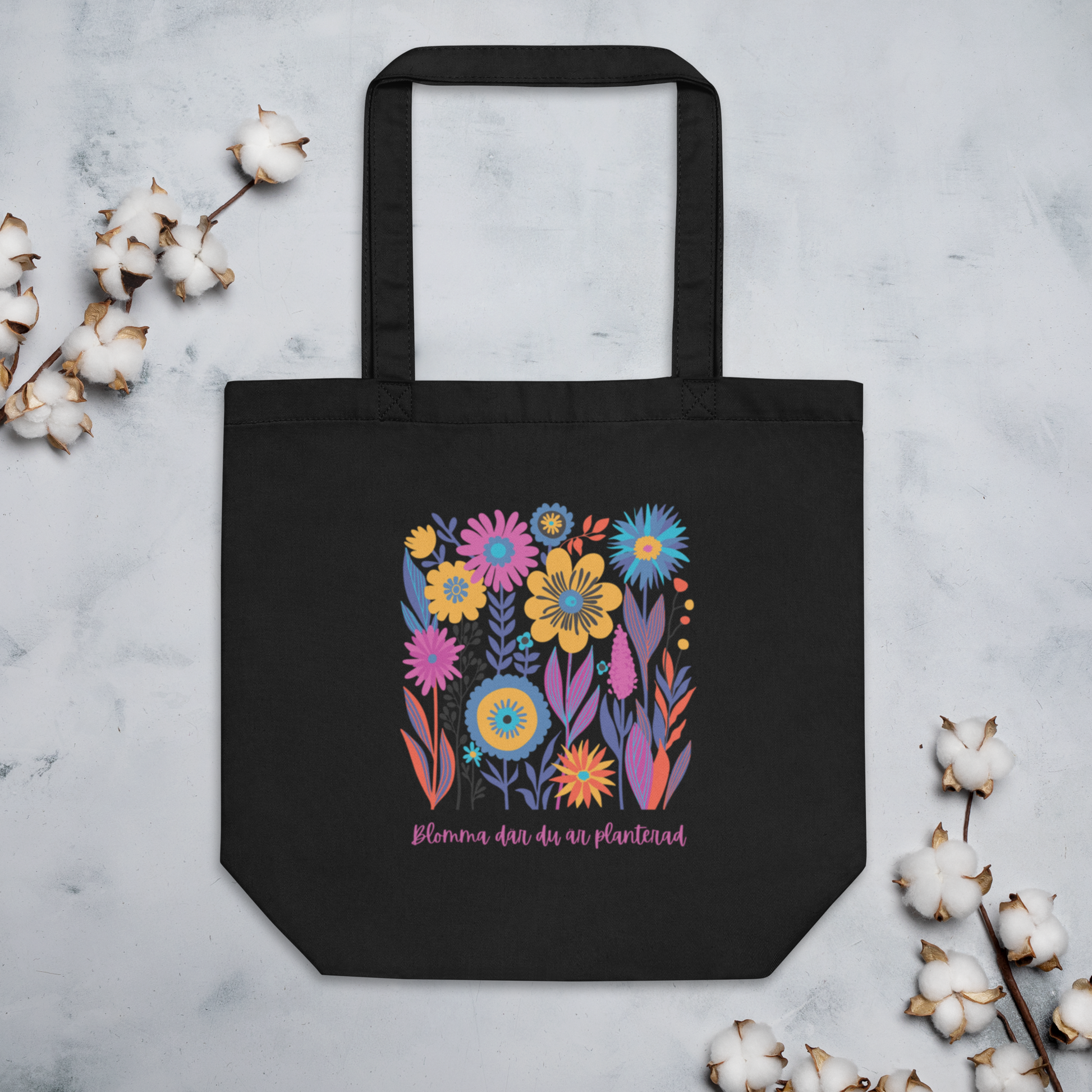 Svart ekologisk tygväska med färgglada illustrerade blommor och texten "Blomma där du är planterad" i rosa.
Black organic tote bag with colorful illustrated flowers and the text "Blomma där du är planterad" in pink.