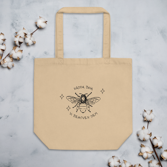 Naturfärgad beige ekologisk tygväska med svart handritat bi och texten "Rädda bina – vi behöver dem" runtom.
Natural-colored oyster organic tote bag with a black hand-drawn bee and the text "Rädda bina – vi behöver dem" surrounding it.