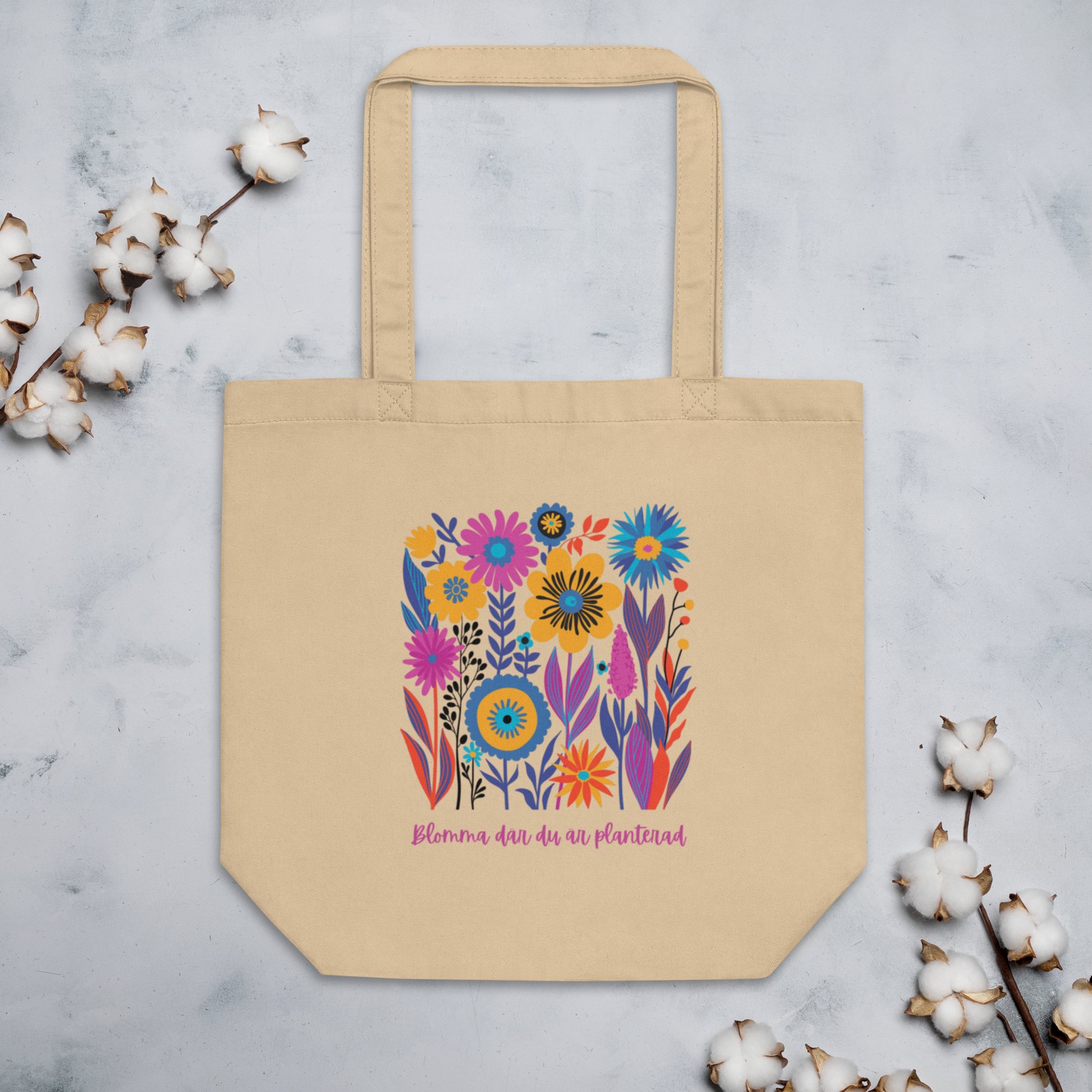 Naturfärgad beige ekologisk tygväska med färgglada illustrerade blommor och texten "Blomma där du är planterad" i rosa.
Natural-colored oyster organic tote bag with colorful illustrated flowers and the text "Blomma där du är planterad" in pink.