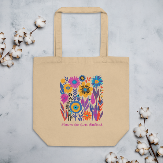 Naturfärgad beige ekologisk tygväska med färgglada illustrerade blommor och texten "Blomma där du är planterad" i rosa.
Natural-colored oyster organic tote bag with colorful illustrated flowers and the text "Blomma där du är planterad" in pink.